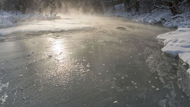 Glittering frozen river reflecting low sun with rising mist and snow-covered banks, winter