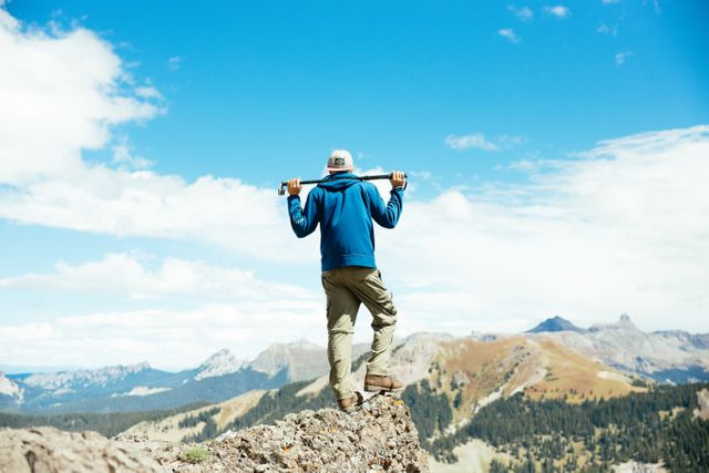 Hiker standing on rocky peak, holding a pole across shoulders, taking in expansive mountain view. Ideal for illustrating themes of adventure, outdoor activities, travel, and exploration. Perfect for websites, travel blogs, brochures, and advertisements focused on nature, hiking, adventure tours, and healthy lifestyles.