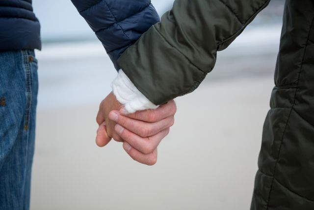 Close-up of a couple holding hands on a beach, showcasing their connection and affection. Ideal for use in articles or advertisements about relationships, love, and romantic getaways. Can also be used in social media posts or blogs focusing on couple activities, bonding, and emotional support.