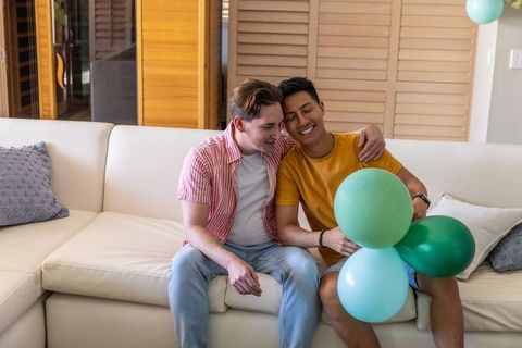 Diverse male couple sitting on cream sofa holding green balloons celebrating home moment
