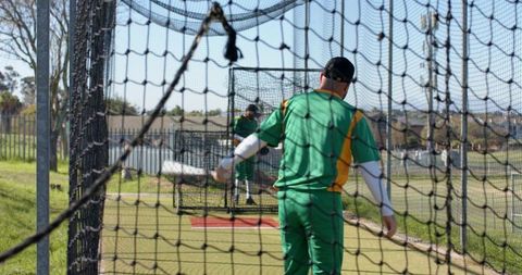 Baseball teammates practicing batting in netted cage wearing green yellow uniforms outdoor training
