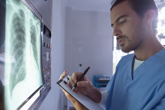 Male surgeon writing on clipboard while examining x-ray on x-ray light box in operation room at hospital