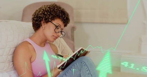 Young woman journaling and tracking finance data at home with rising green graph overlay