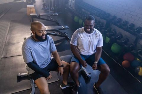 African american men laughing and resting after strength training on benches near dumbbells in gym