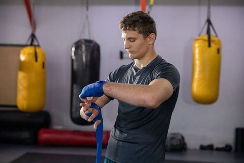 Male athlete wrapping hands with blue wraps in boxing gym with hanging heavy bags
