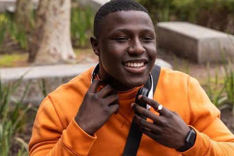 Smiling cheerful african american man in orange crewneck adjusting headphones on park bench
