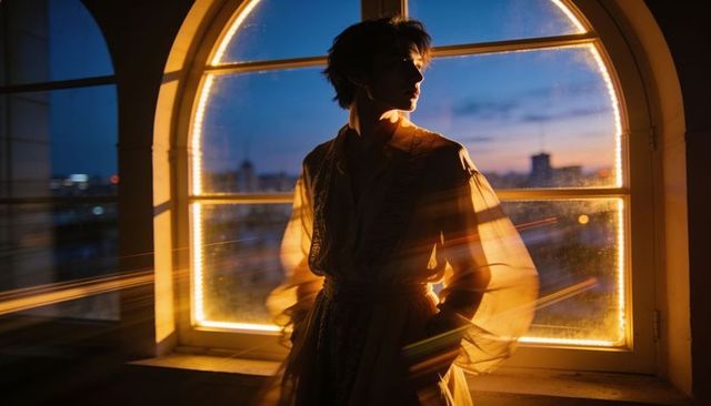 Silhouetted woman standing by arched led-lit window at dusk, cinematic rimelight over city