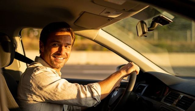 Smiling adult man driving modern car at golden hour gripping steering wheel with dashcam