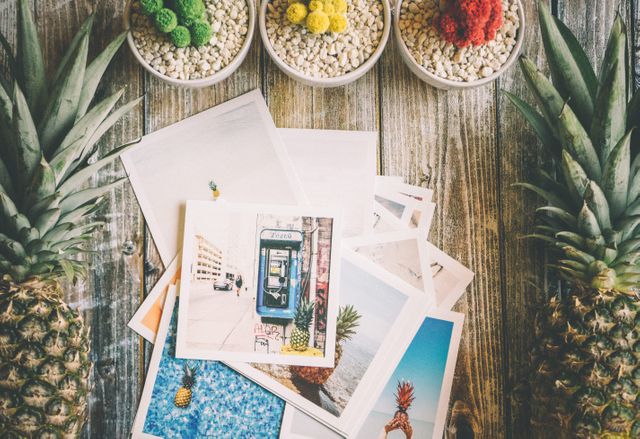Colorful travel photos arranged on wooden table, accompanied by pineapples and decorative succulent plants, evoke sense of summer adventure and tropical getaway. Perfect for travel blogs, vacation planning materials, tourism brochures, and social media posts celebrating summer.