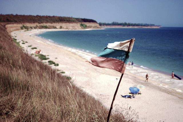 A weathered flag stands on a cliff of grass, overlooking a secluded sandy beach with a few people enjoying the summer day. The calm sea meets the horizon, making it an ideal image for use in travel brochures, summer vacation advertisements, and websites promoting outdoor activities, nature escapes, or peaceful retreats.