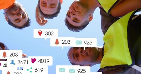 Overhead team huddle with digital notifications and high-visibility vests under blue sky