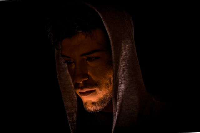 Young man wears hoodie, face partially illuminated by low light, background in darkness. Image evokes brooding and introspection. Suitable for themes around contemplation, solitude, mystery, and dark moods. Ideal for illustrating blogs, articles, or artistic projects that focus on serious emotions or introspective topics.