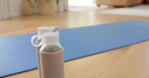 Reusable water bottles beside blue yoga mat on sunlit wood floor promoting home wellness