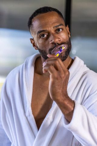 Mature african man brushing teeth in white bathrobe, morning grooming portrait
