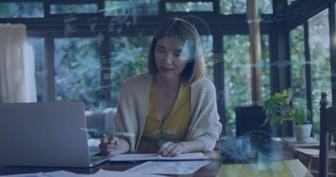 Working from home: woman reviewing documents with laptop and pen in sunlit workspace