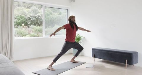 African American man practicing yoga at home on mat with laptop in bright minimalist living room