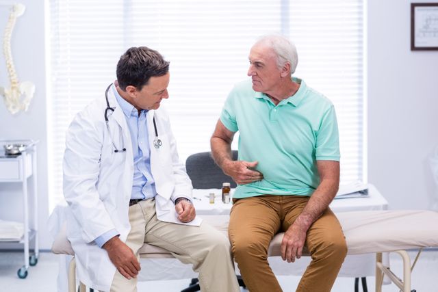 Senior man sitting on examination table, showing stomach pain to doctor in clinic. Doctor attentively listening and assessing patient's condition. Useful for healthcare, medical consultation, elderly care, and patient-doctor interaction themes.