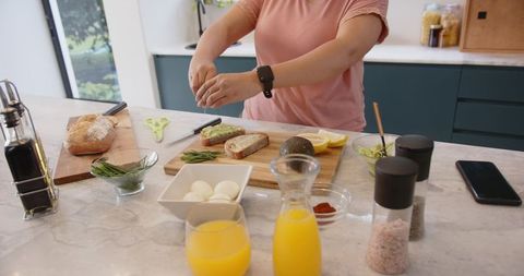 Woman preparing avocado toast on marble counter with fresh herbs and orange juice