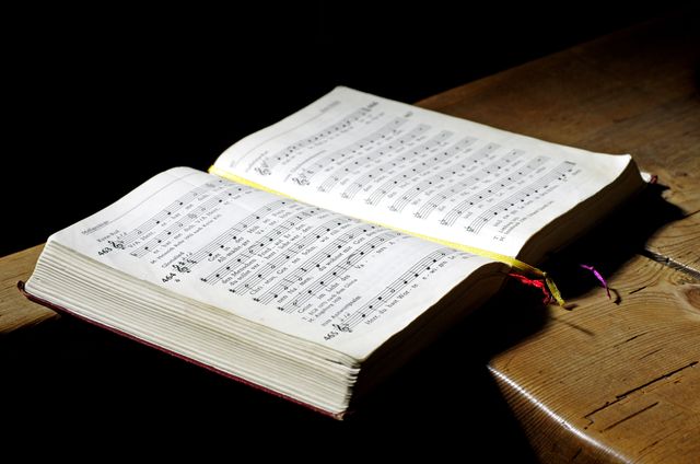 Open book of sheet music rests on wooden desk in dim lighting. Suitable for articles and blogs about music education, classical music, studying music notation, hymnals, or book reviews related to music.