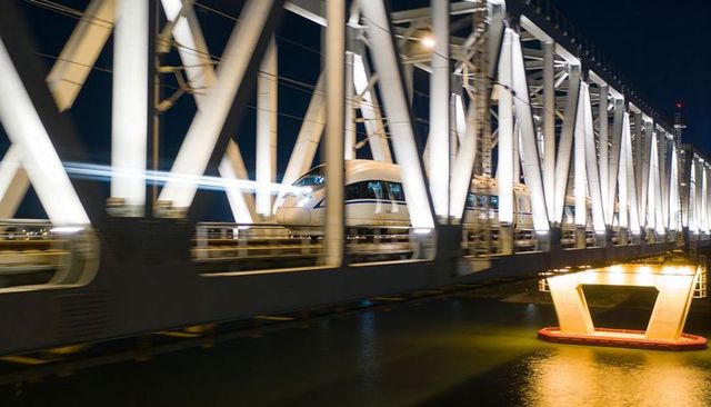 Racing bullet train crossing illuminated steel truss bridge at night with river reflections