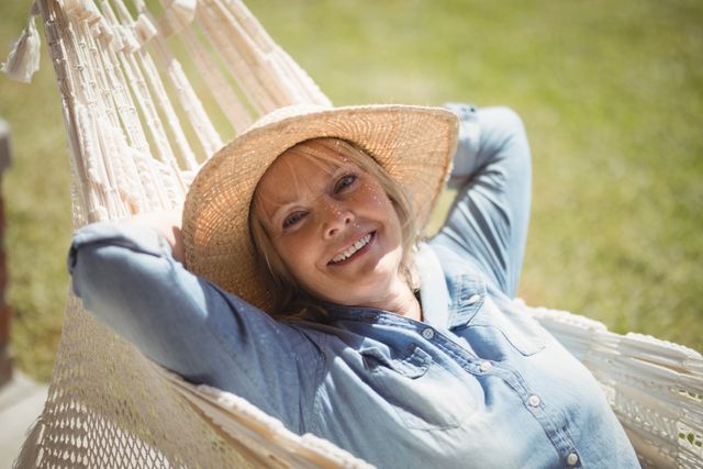 Senior woman enjoying a sunny day while relaxing on a hammock. She is smiling and wearing a casual outfit with a hat. Ideal for use in advertisements promoting retirement, leisure activities, outdoor lifestyle, and senior well-being.