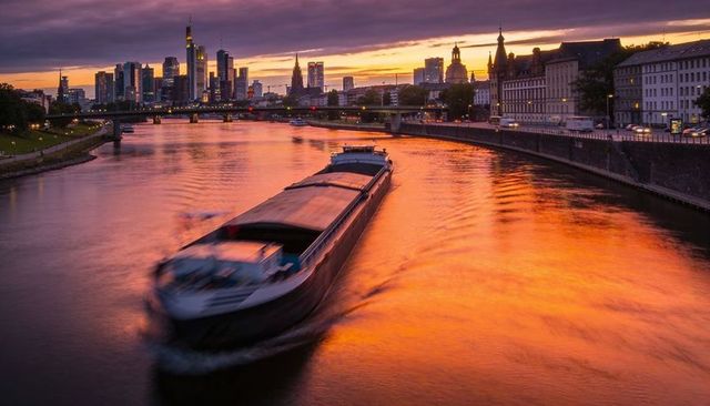 Gliding cargo barge at sunset along river reflecting city skyline and golden promenade