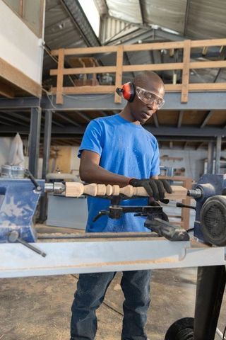 African american man operating wood lathe wearing safety gear in busy carpentry workshop