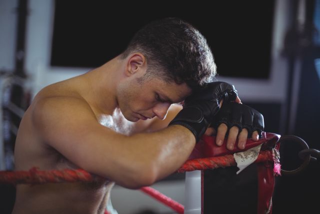 Boxer leaning on ropes of boxing ring, showing signs of exhaustion and determination. Ideal for use in sports and fitness articles, motivational content, training programs, and advertisements for athletic gear.
