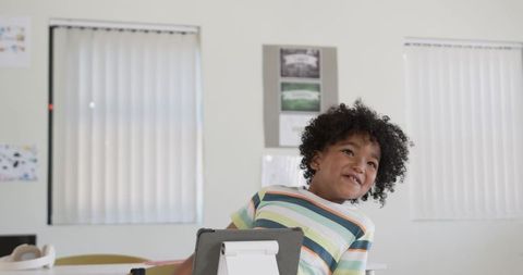 Curly-haired African American boy using tablet and headset in classroom, smiling while learning