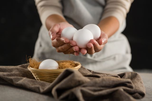 Mid-section of woman holding white eggs in hands, with a basket of eggs on a rustic cloth. Ideal for use in cooking blogs, organic food promotions, kitchen-related advertisements, and culinary magazines.