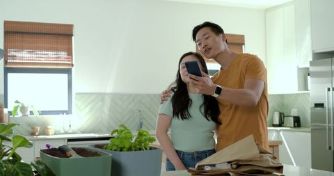 Asian couple planning indoor herb garden in sunlit modern kitchen using smartphone