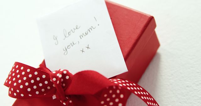 A red gift box adorned with a polka-dotted ribbon and a note expressing love for mom, with copy space. Such a present is often associated with Mother's Day, birthdays, or as a token of appreciation.