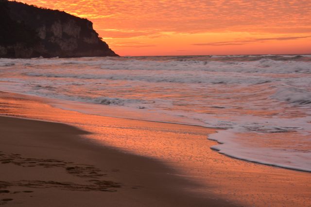 A stunning sunset over a peaceful beach with gentle waves washing the shore. The sky is painted with brilliant orange hues, reflecting on the water’s surface. Perfect for use in travel websites, relaxation and meditation materials, and marketing campaigns focusing on tranquil destinations.