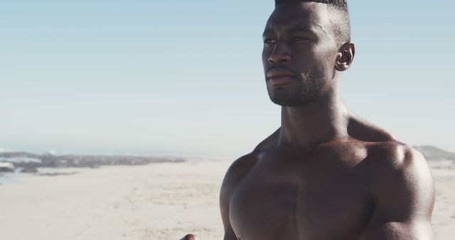Young athletic man standing by beach, looking into distance with determined expression. Muscular build signifies fitness and strength, ideal for promoting active lifestyle, sports brands, or health and wellness campaigns. Beach and ocean backdrop suitable for travel websites or nature-focused advertisements.
