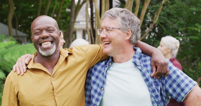 Two senior men are enjoying a lighthearted moment together, with their arms around each other and vibrant smiles on their faces. They appear relaxed and cheerful, standing in a lush green outdoor setting. This image can be used to convey themes of friendship, joy, and positive aging, and is great for use in health and lifestyle promotions, senior living advertisements, lifestyle blogs, or wellness websites.