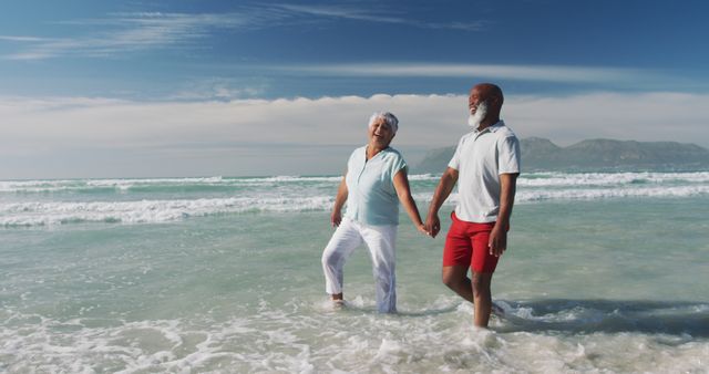 Senior couple is walking hand in hand along sunny beach with ocean waves rolling in. This can be used in advertisements for vacation companies, retirement plans, healthcare, or lifestyle articles focused on senior life and relationships.