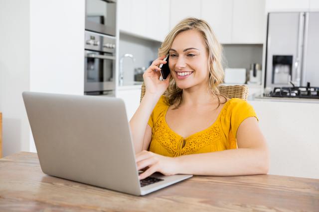 Woman in a yellow top talking on a mobile phone while using a laptop in a modern kitchen. Ideal for illustrating remote work, home office setups, modern lifestyles, and technology use in everyday life.