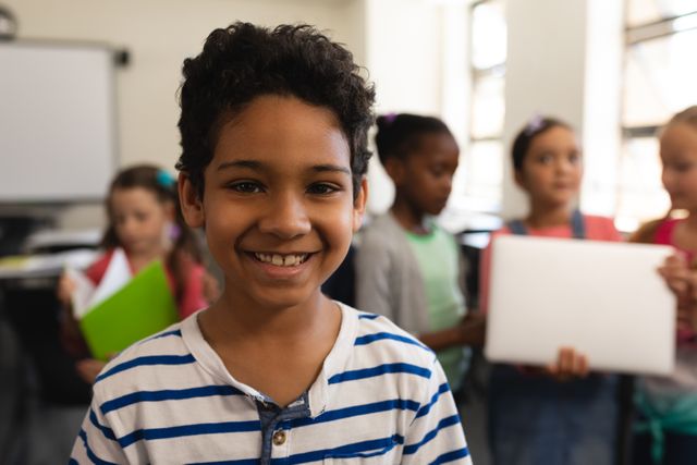 Close-up of a happy schoolboy smiling at the camera in a classroom setting. Other children are in the background, holding books and engaging with each other. Perfect for educational materials, school advertisements, and articles on childhood education and diversity.
