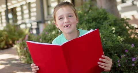 Schoolgirl reading aloud with bright red folder in sunlit courtyard