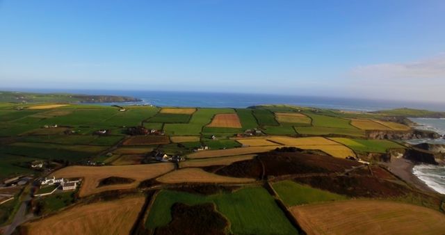 Aerial view showcases a picturesque rural landscape with patchwork fields leading to a coastal area, under a clear blue sky. The image captures the serene beauty of the countryside meeting the ocean.