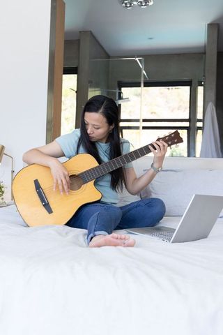 Mature woman practicing acoustic guitar on bed while following online lesson on laptop