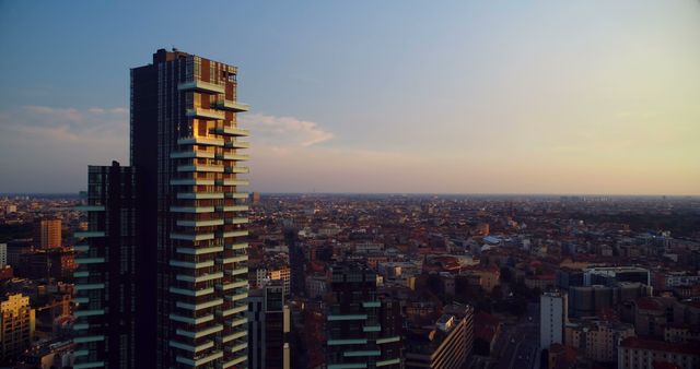 Sunset scene over a bustling cityscape with a prominent modern high-rise building in the foreground. The view showcases urban development and residential spaces, perfect for use in real estate advertising, travel promotions, and urban planning presentations.