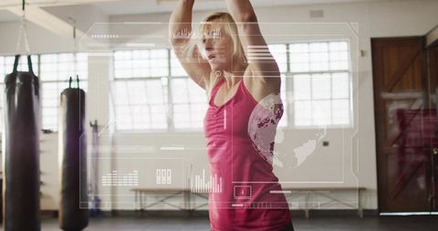 Mid-adult woman training, raising arms overhead with hud fitness data in boxing gym