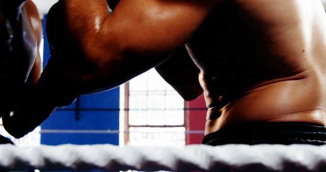 A close-up view captures the muscular torso of a male boxer in the ring, highlighting his athletic form and strength. His intense training and dedication to the sport are evident in the defined muscles and the boxing environment around him.