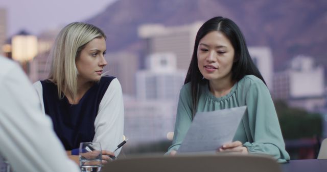 Image of two diverse female colleagues discussing document at evening office meeting. Business, communication, inclusivity and flexible working concept digitally generated image.