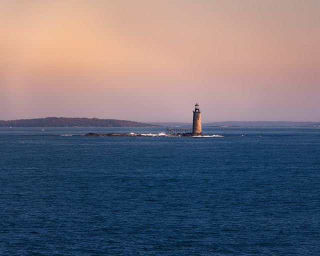 Solitary lighthouse stands amid calm blue sea with pinkish-orange hues in sky at sunset. Useful for travel websites, coastal tourism promotions, and maritime themed artwork or desktop backgrounds. Highlights tranquility and natural beauty of coastal areas.