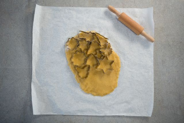This image shows a top view of rolled dough with various shaped pastry cutters on wax paper, accompanied by a rolling pin. Ideal for use in baking blogs, recipe websites, cooking tutorials, and kitchen-themed advertisements.