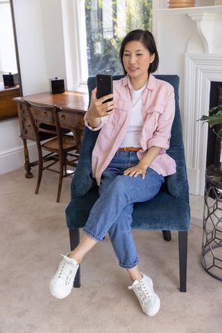 Asian woman in late 20s sitting in blue chair using smartphone at home desk casual modern cozy