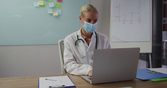 Caucasian female doctor wearing mask sitting at desk in meeting room using laptop. medical professionals at work during covid 19 coronavirus pandemic.