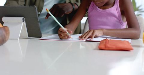 African american father guiding daughter doing homework at table with tablet